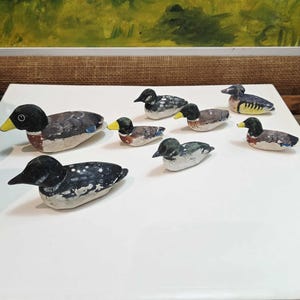 Puede incluir: Una colección de figuras de patos de madera pintadas a mano. Los patos presentan plumaje negro, blanco, marrón y azul con picos amarillos. Las figuras están dispuestas sobre una superficie blanca, con una pintura borrosa en el fondo.