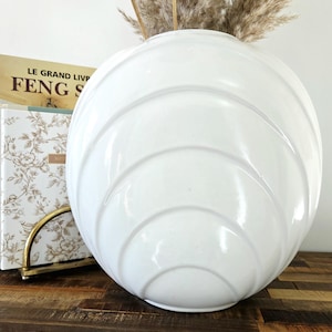 Peut inclure: Un grand vase en céramique blanc, de forme arrondie, orné d'une série de rainures incurvées en forme de vagues. Le vase est posé sur une surface en bois, avec un livre et des plantes séchées en arrière-plan. Le texte "LE GRAND LIVRE" et "FENG SHUI" est visible.