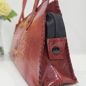 May include: A reddish-brown leather handbag with a stitched design and a black zipper closure. The bag has two handles and a decorative floral pattern. The leather has a glossy finish and the stitching is visible along the edges.