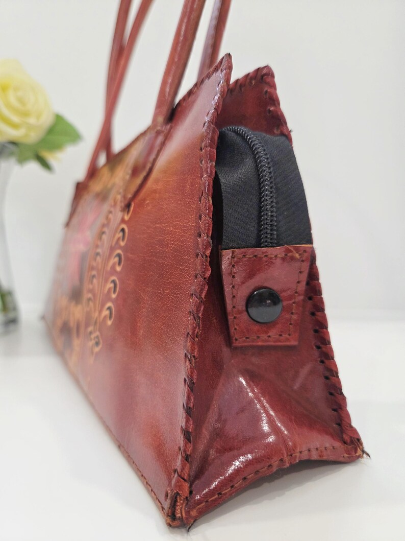 May include: A reddish-brown leather handbag with a stitched design and a black zipper closure. The bag has two handles and a decorative floral pattern. The leather has a glossy finish and the stitching is visible along the edges.