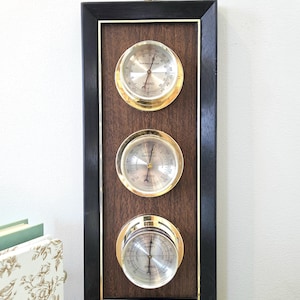 Puede incluir: Una estación meteorológica vintage con marco de madera oscura y detalles en latón. Cuenta con un termómetro, un barómetro y un higrómetro. Los instrumentos tienen esferas plateadas y están montados en un panel de madera oscura. Un asa de latón en la parte superior.