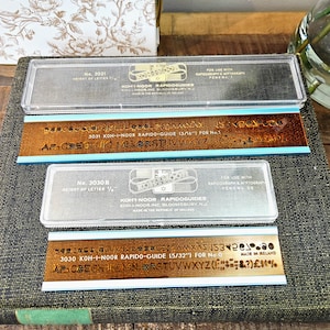 Pode incluir: Dois Rapidoguides Koh-I-Noor, modelos 3031 e 3030B, com estojos de plástico transparente. Os guias são castanhos com detalhes azuis e apresentam guias de letras para canetas. A altura das letras é de 1,11 e 1,19 cm.