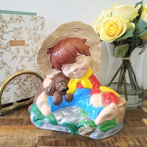 Peut inclure: Figurine en céramique représentant un jeune garçon aux cheveux bruns, une écharpe jaune et une salopette bleue, étreignant un chien marron. Le garçon tient une pomme. La figurine est sur une base avec des accents verts et gris.
