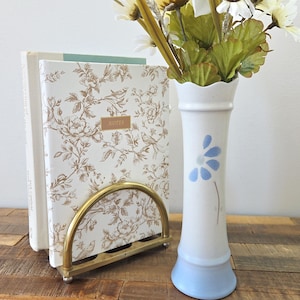 Peut inclure: Un vase blanc avec un motif floral bleu et une base bleu clair contient des fleurs artificielles. Un porte-serviettes en métal doré soutient deux carnets à motifs floraux. L'un des carnets porte une étiquette dorée avec le mot "NOTES".
