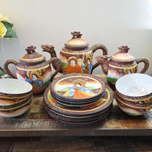Op de afbeelding: Een keramische theeset met twee theepotten en één grotere theepot, elk met een draakvormig handvat en dekselknop. De set bevat bijpassende borden en kommen versierd met kleurrijke figuren in traditionele kleding tegen een donkerbruine achtergrond.