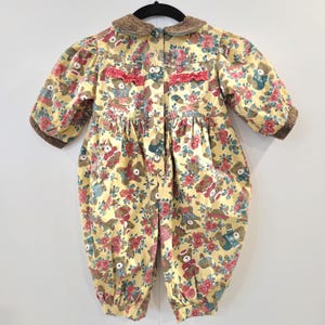 May include: A yellow baby romper with a floral and teddy bear pattern. The romper has a brown collar and cuffs, with red accents and buttons down the front. The fabric is a soft, patterned material.