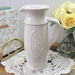 May include: A cream-colored ceramic pitcher with a handle and spout. The pitcher has an embossed design of a person playing a harp. The pitcher is sitting on a white lace doily.