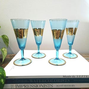 May include: Four vintage champagne flutes. The glasses are light blue with gold accents. The base and rim of each glass are trimmed in gold. The center of each glass has a gold textured design. The glasses are on a white surface.