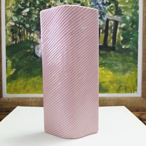 May include: A rectangular, light pink ceramic vase with a textured, diagonal striped pattern. The vase is displayed on a white surface, with a framed painting in the background.
