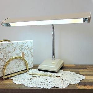 Puede incluir: Una lámpara de escritorio beige con cabezal rectangular y cuello cromado flexible. Una caja con estampado floral y un bolígrafo dorado con extremo perlado están sobre una superficie de madera. También hay un tapete de encaje blanco.