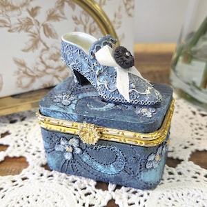Peut inclure: Une boîte à bijoux décorative bleue avec une chaussure à talon assortie sur le dessus. La boîte a une bordure dorée et des motifs floraux. La chaussure a un nœud blanc et un bouton noir.