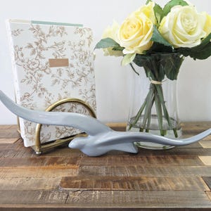 Op de afbeelding: Een lichtgrijze keramische vogelsculptuur in vlucht, geplaatst op een houten oppervlak. Een boek met bloemmotief en een boeket gele rozen in een glazen vaas staan op de achtergrond. De vogelsculptuur is ongeveer 30 cm lang.