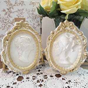 May include: Two white oval cameo plaques with gold trim. Each plaque features a profile of a woman. The frames have ornate gold detailing. The background includes a floral patterned notebook and a vase of yellow roses.