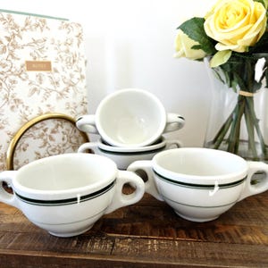 Peut inclure: Ensemble de tasses à soupe en céramique blanche, avec une bande verte foncée sur le bord et une anse. Les tasses sont empilées et disposées sur une surface en bois. Un carnet floral et un vase de roses jaunes sont en arrière-plan.