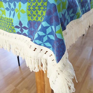 Peut inclure: Nappe colorée avec un motif géométrique dans les tons de bleu, vert et violet. La nappe est ornée d'une frange blanche sur les bords. Le tissu est drapé sur une table en bois, créant un aspect décoratif et texturé.