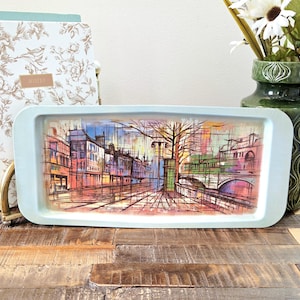 Puede incluir: Una bandeja rectangular de color azul claro que presenta una ilustración artística abstracta y colorida de una escena de calle urbana con edificios, un puente y árboles desnudos. La obra utiliza una mezcla de líneas naranjas, rosas, verdes y negras.