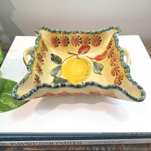 May include: A square ceramic serving bowl with a yellow base and a scalloped edge. The bowl is decorated with a hand-painted lemon and floral design. The rim is trimmed with a green and red pattern. It has two small handles.