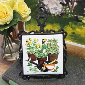 Peut inclure: Un dessous de plat en métal noir avec un carreau de céramique carré représentant une illustration botanique d'herbes en pots, bouteilles et un mortier et un pilon. Le dessous de plat a un cadre décoratif et une poignée. Roses jaunes dans un vase en arrière-plan.