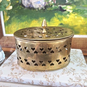 Op de afbeelding: Een vintage messing container met een deksel met hartvormige uitsparingen en een decoratieve bekroning. De ronde container heeft een geperforeerde bovenkant en wordt waarschijnlijk gebruikt voor opslag of weergave. Het messing heeft een warme, verouderde patina.