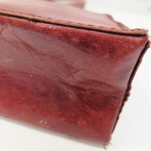 May include: Close-up of a burgundy leather handbag. The bag has a structured shape with visible stitching along the edges. The leather shows signs of wear, adding to its vintage appeal. The handle is visible in the background.