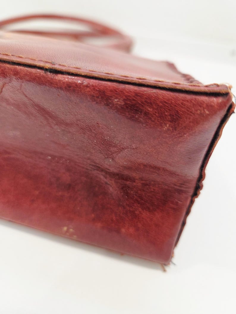 May include: Close-up of a burgundy leather handbag. The bag has a structured shape with visible stitching along the edges. The leather shows signs of wear, adding to its vintage appeal. The handle is visible in the background.