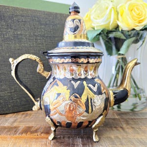 May include: A decorative vintage teapot with a black body, gold detailing, and detailed metalwork. The teapot has a curved handle, a long spout, and a decorative lid. The design incorporates figures and patterns in gold, silver, and copper tones. It rests on three small feet.