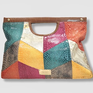 Peut inclure: Une pochette patchwork multicolore avec une poignée et une bordure marron. Le sac présente une variété de textures et de couleurs, notamment le turquoise, l'argent, l'orange et le jaune. Une petite plaque dorée avec le nom de la marque est visible.