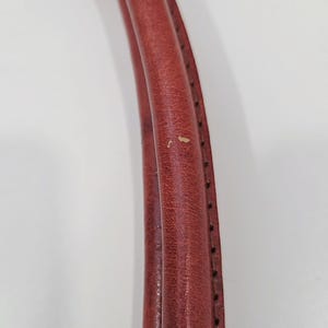 May include: Close-up of a pair of red leather bag handles. The handles are curved and stitched with black thread along the edges. The leather shows signs of wear with small imperfections.