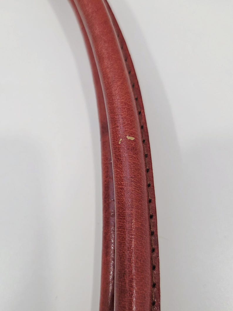 May include: Close-up of a pair of red leather bag handles. The handles are curved and stitched with black thread along the edges. The leather shows signs of wear with small imperfections.