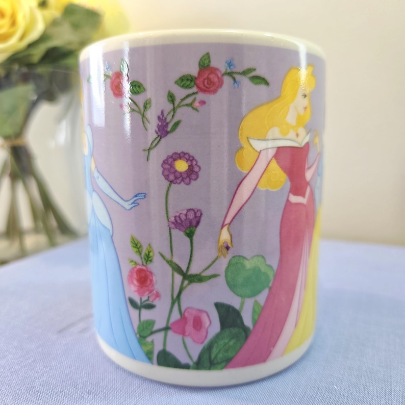 Op de afbeelding: Een cilindrische keramische mok met illustraties van animatiefiguren en bloemmotieven. De mok heeft een lavendelkleurige achtergrond met afbeeldingen van een prinses in een roze jurk en een prinses in een blauwe jurk, omringd door bloemen.
