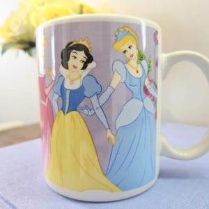 Op de afbeelding: Witte keramische mok met illustraties van geanimeerde prinsessen in kleurrijke jurken. De mok heeft een gebogen handvat en een lichtpaarse achtergrond met bloemmotieven. De prinsessen zijn Sneeuwwitje en Assepoester.