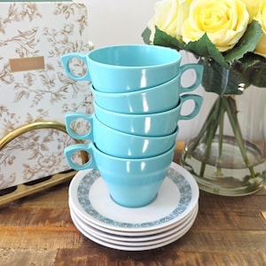 May include: Five turquoise ceramic teacups with distinctive handles, stacked on matching saucers with a blue and white design. The cups are on a wooden surface, with a floral notebook and yellow roses in the background. The cups are approximately 9 cm in diameter.