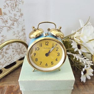 May include: A vintage blue and gold alarm clock with a yellow face, featuring black hands and Arabic numerals. The clock, labeled "Alpine," sits on a light green box. The clock has a gold-colored frame and bells. The clock is Made in Germany.