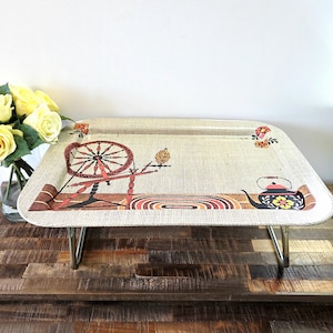 Peut inclure: Un plateau de service beige vintage avec des pieds pliants en métal. Le plateau présente une impression décorative d'un rouet, d'une théière et d'éléments floraux dans des tons de rouge, de marron et d'orange. Le plateau est rectangulaire avec des coins arrondis.