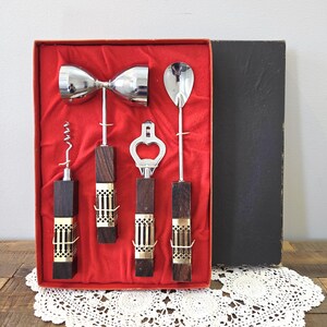 Può includere: Un set di attrezzi da bar vintage in una scatola rossa. Il set include un jigger doppio, un cavatappi, un apribottiglie e un cucchiaio da bar. Gli attrezzi hanno manici in legno scuro con dettagli in metallo dorato.