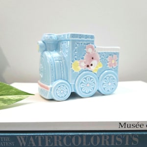 Peut inclure: Un pot de fleurs en céramique bleu clair en forme de train avec un motif d'ours en peluche rose et jaune. Le train a trois roues avec des détails floraux et un accent rose et blanc. Le pot est posé sur une surface blanche.