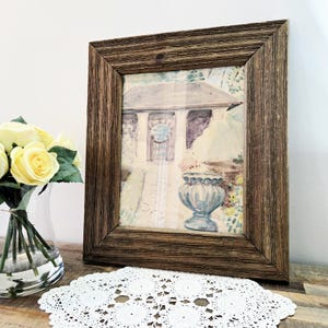 Peut inclure: Une peinture à l'aquarelle encadrée représentant une scène de jardin avec une structure en pierre et une urne bleue. Le cadre est en bois brun foncé et rustique. Un vase de roses jaunes et un napperon en dentelle blanche sont au premier plan.