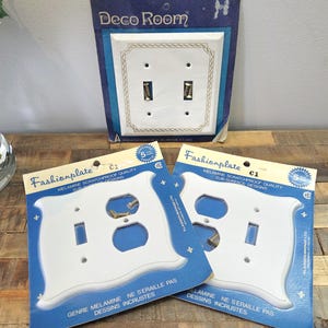 May include: White melamine switch plates with gold-toned accents and hardware, packaged in blue and white cardboard. One package reads "Deco Room", while others say "Fashionplate". The plates are for light switches and electrical outlets.