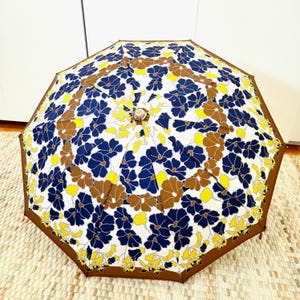 Pu&ograve; includere: Un ombrello aperto con un motivo floreale composto da fiori blu navy, marroni e gialli su sfondo bianco. Ha un bordo marrone e una parte superiore decorativa. L'ombrello &egrave; appoggiato su un tappeto dai colori neutri.