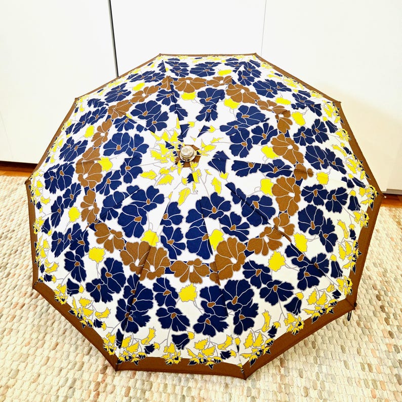 Pu&ograve; includere: Un ombrello aperto con un motivo floreale composto da fiori blu navy, marroni e gialli su sfondo bianco. Ha un bordo marrone e una parte superiore decorativa. L'ombrello &egrave; appoggiato su un tappeto dai colori neutri.
