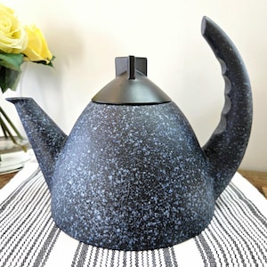 May include: A speckled black and blue ceramic teapot with a curved handle and a spout. The teapot has a black lid and sits on a striped white and black cloth. A vase of yellow roses is in the background.