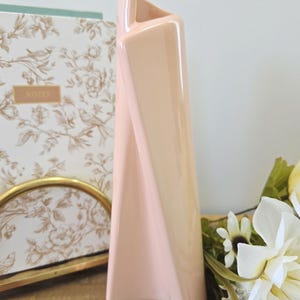 Peut inclure: Un vase en céramique pêche de forme haute et torsadée. Il présente une finition brillante et une forme rectangulaire qui s'incurve et s'effile vers le haut. Le vase est posé sur une surface en bois, à côté d'un carnet floral et d'un cadre doré.