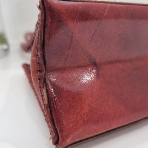 May include: Close-up of a burgundy leather bag. The bag's corner is visible, showcasing the stitched seams and the texture of the leather. The leather has a glossy finish and shows signs of wear.