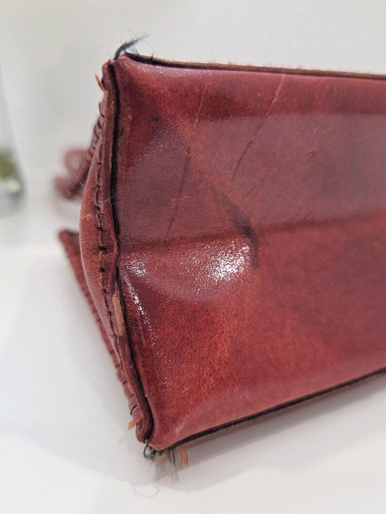May include: Close-up of a burgundy leather bag. The bag's corner is visible, showcasing the stitched seams and the texture of the leather. The leather has a glossy finish and shows signs of wear.