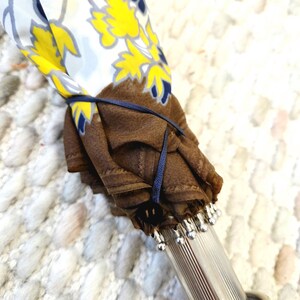 Pu&ograve; includere: Un ombrello chiuso con un motivo floreale giallo, bianco e blu. Il tessuto dell'ombrello &egrave; fissato con un nastro blu navy. L'impugnatura &egrave; argentata con perline decorative argentate.