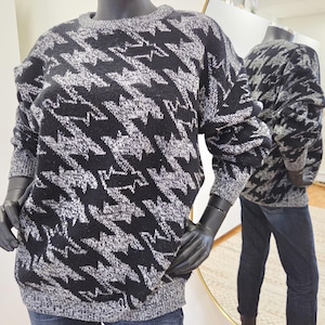 May include: A black and gray houndstooth pattern sweater. The sweater has a crew neck and long sleeves. The sweater is displayed on a mannequin with black gloves and blue jeans. The sweater is reflected in a gold-framed mirror.