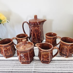 Peut inclure: Un ensemble à café vintage en céramique marron comprenant une cafetière, une crémière, un sucrier avec couvercle et six tasses. Chaque pièce est ornée d'un motif floral. L'ensemble est disposé sur une nappe à rayures.