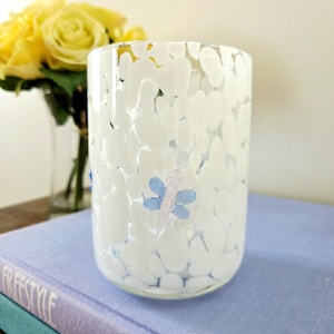 Peut inclure: Un vase en verre transparent avec un motif tacheté blanc et un motif de libellule bleu et rose. Le vase est cylindrique et repose sur une surface bleu clair. Des roses jaunes sont visibles en arrière-plan.