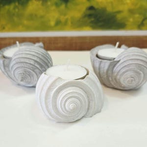 May include: Three seashell-shaped candle holders. Each holder is a light grey colour and has a spiral design. A white tea light candle sits inside each holder. The holders are on a white surface.