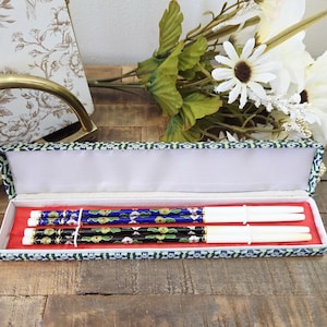 Peut inclure: Un ensemble de baguettes décoratives dans une boîte à motifs. Les baguettes ont des pointes blanches et sont décorées de motifs floraux bleus, noirs et verts. La boîte a un intérieur rouge et un extérieur à motifs. Un bouquet de fleurs blanches est en arrière-plan.